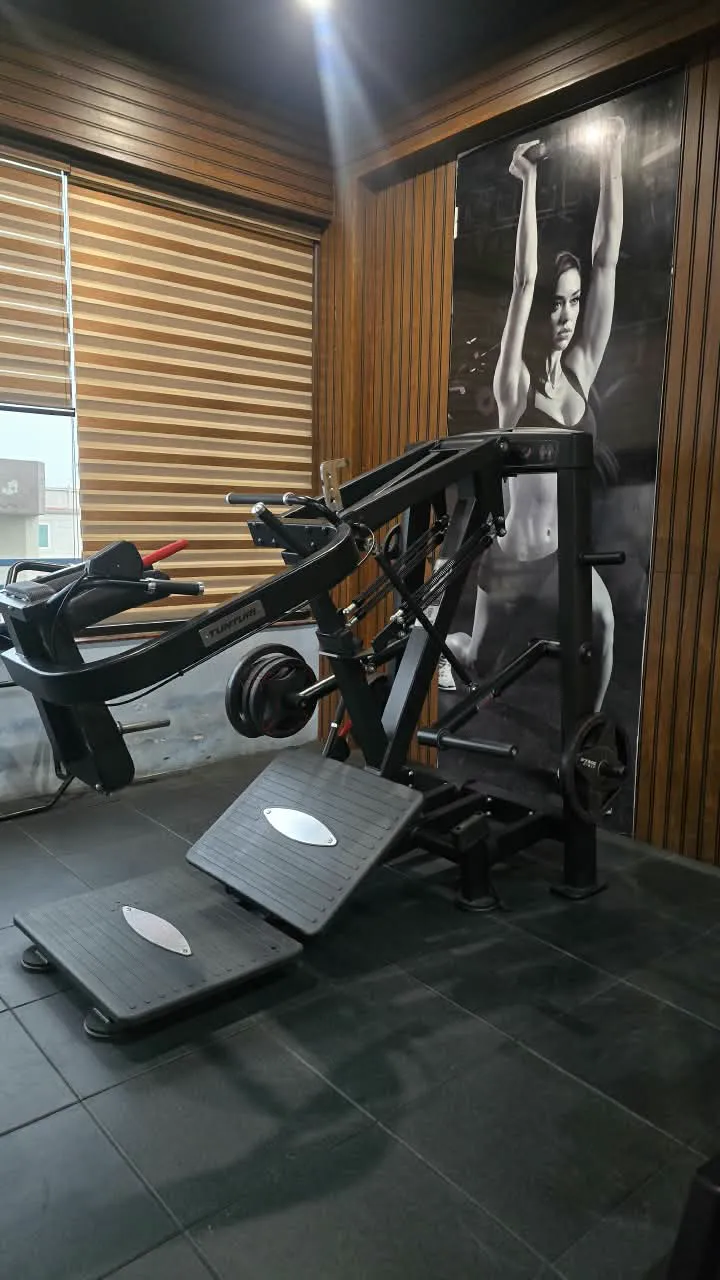 Black plate-loaded leg press with angled footplates against a wood-lined wall and athlete poster